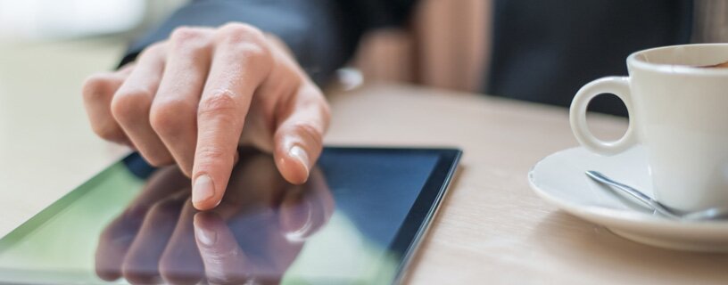 Person bedient ein Tablet mit dem Finger, auf einem Tisch neben einer Tasse Kaffee.